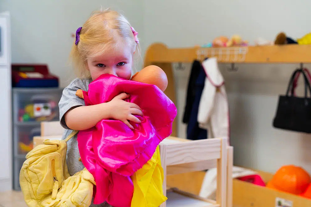 A little girl hugs a doll wrapped in a bright pink blanket, exuding warmth and affection in a playful classroom environment.